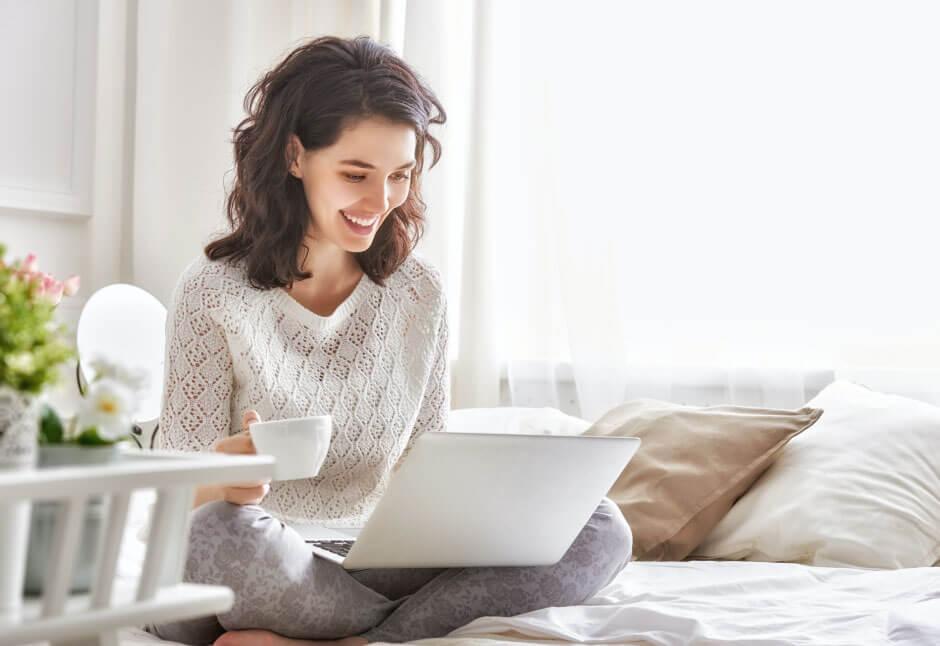 Woman sitting on her bed working with her laptop and with a cup of coffee