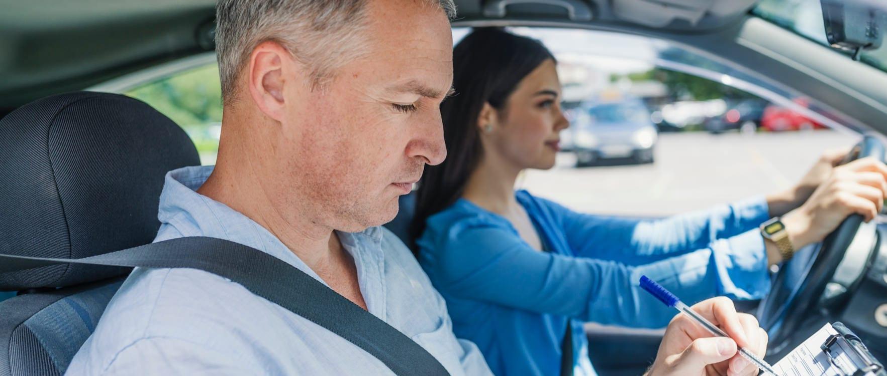 Young woman driving a car with a male instructor sitting at the passenger seat taking notes