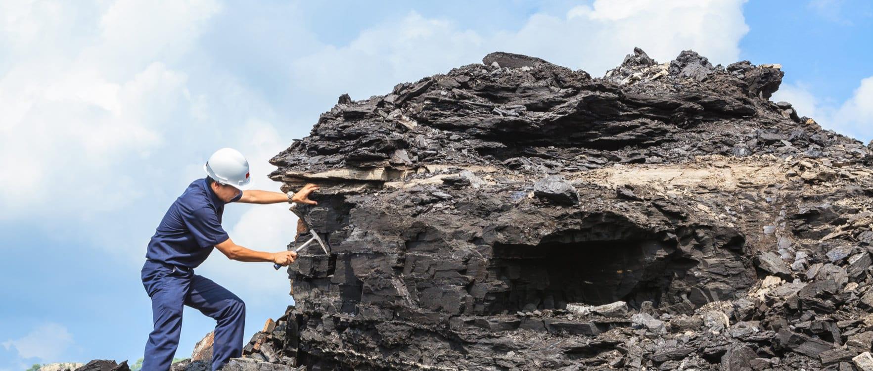 Man with a hard hat and uniform holding a geological hammer standing next to a big rock