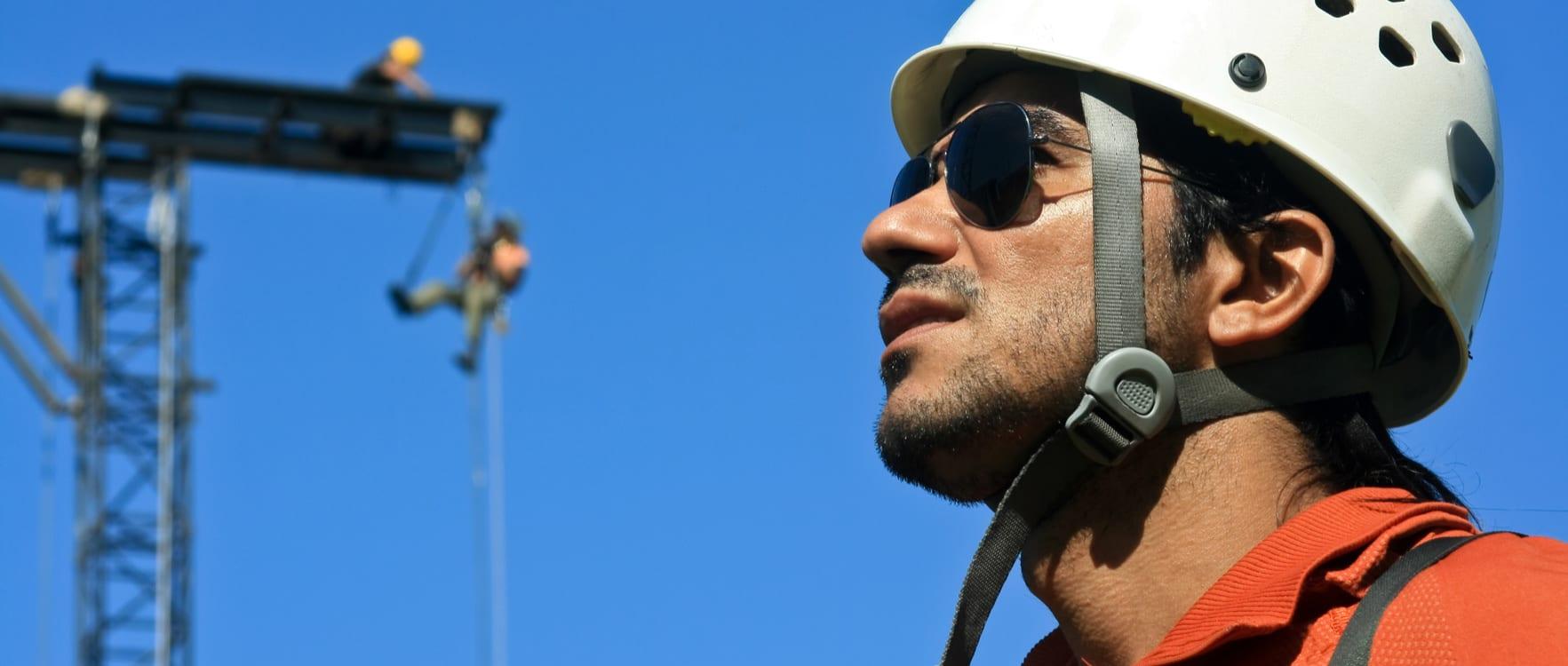 Man with a hard hat and sunglasses with a man rappeling in the background