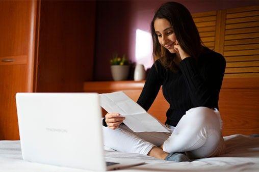 A woman sitting on her bed while holding her resume with her laptop open
